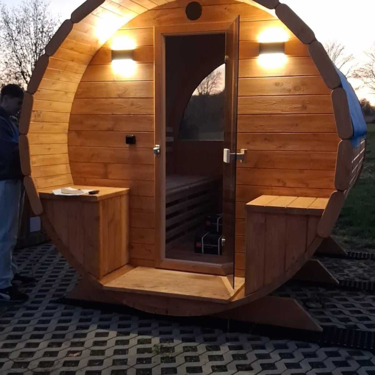 polarsauna shop Fasssauna Highline 285 aus Holz mit Terrasse, Glastür und Außenbeleuchtung im Garten