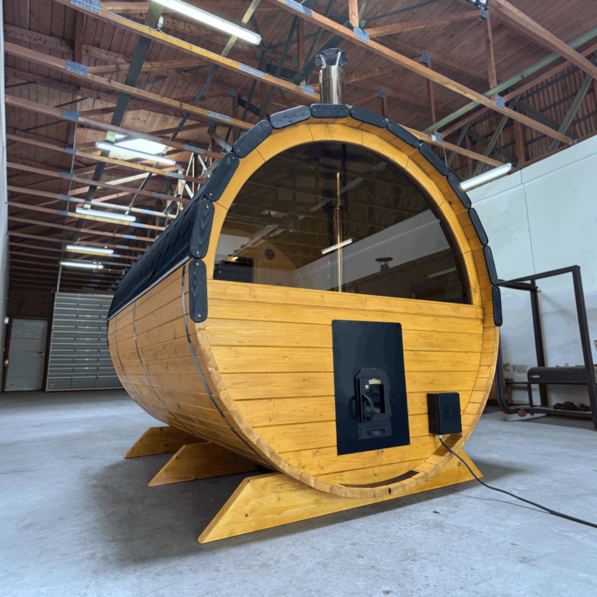 polarsauna shop Fasssauna Highline aus massiver Fichte mit Panorama-Glasfront und Holzofen in einer Lagerhalle