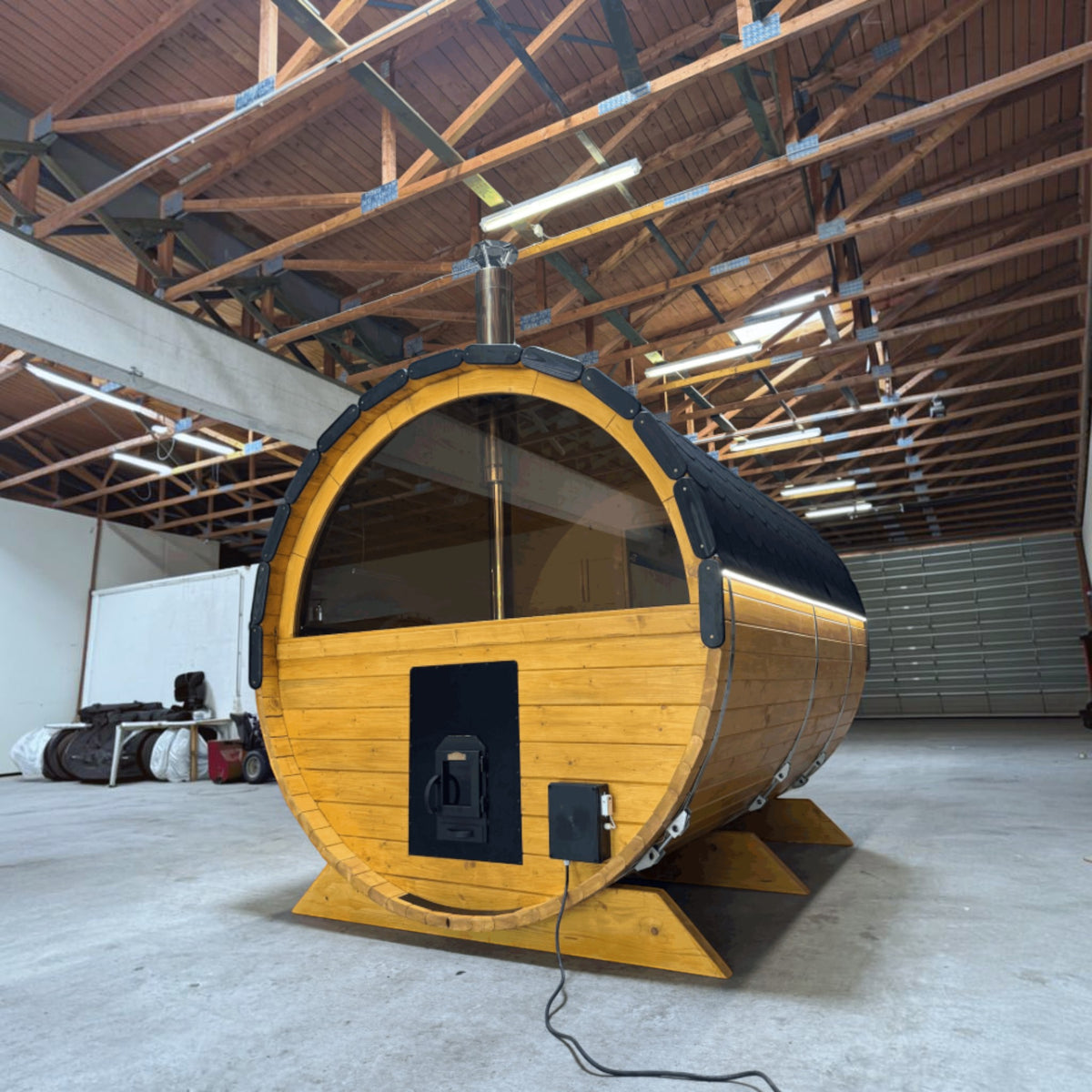 polarsauna shop Fasssauna Highline aus massiver Fichte mit 225 cm Durchmesser und Holzofen in einer Lagerhalle